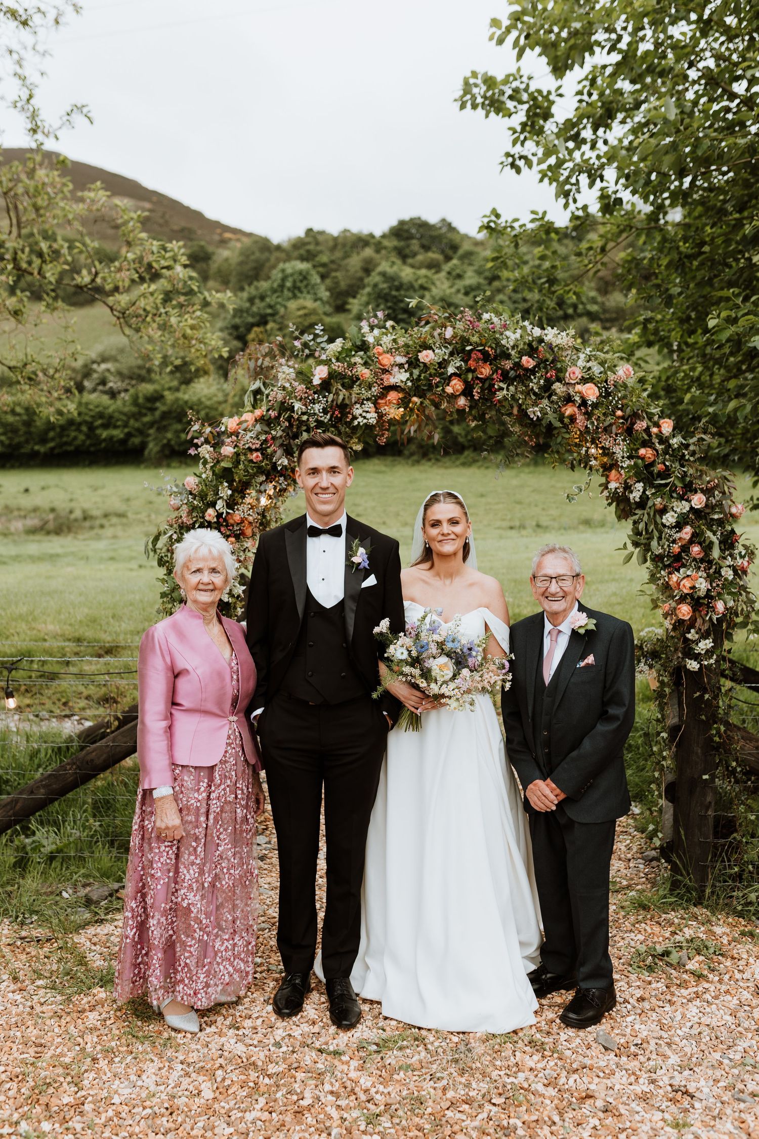 Wedding party poses under floral arch in outdoor garden setting with pink and formal attire.