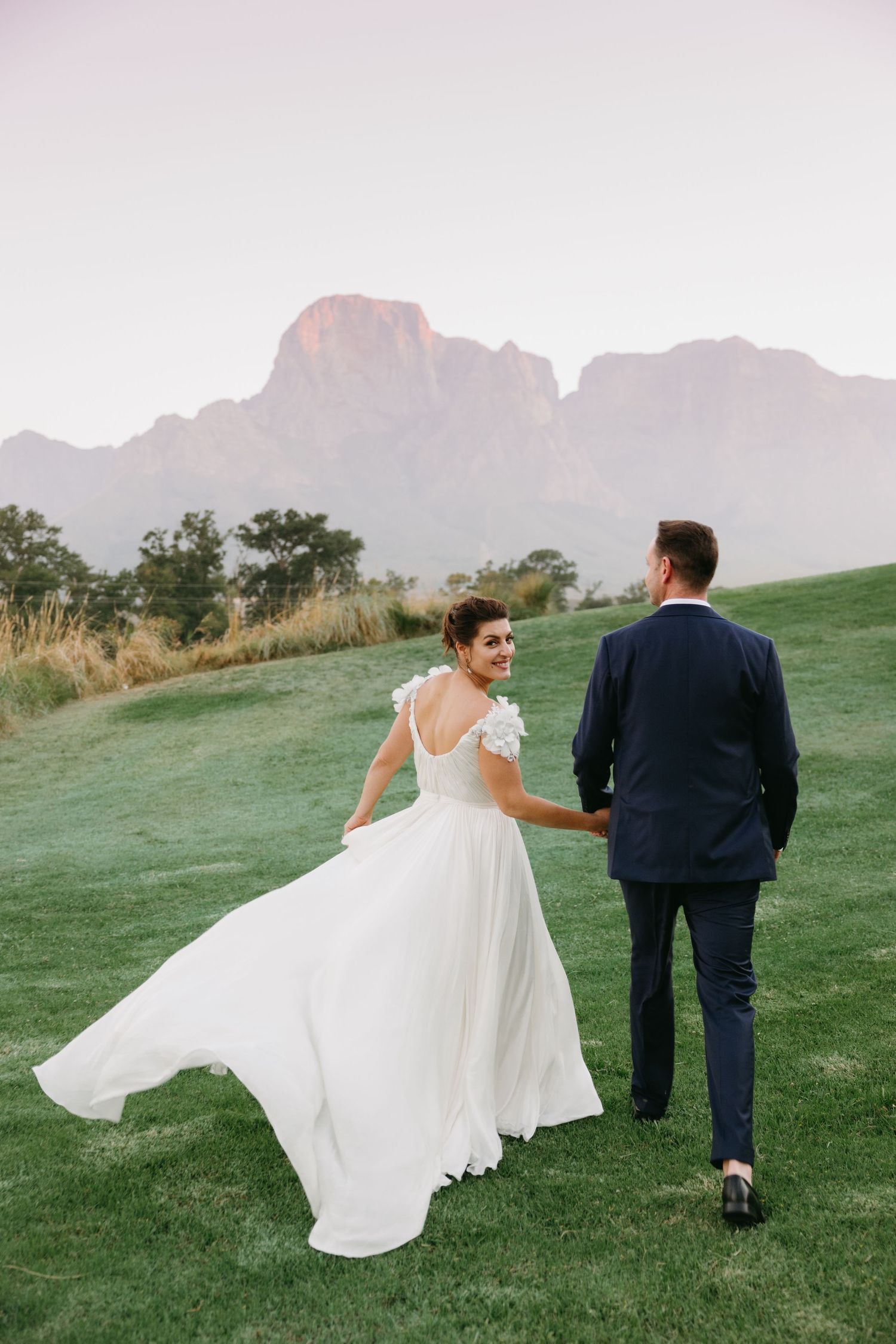 A couple in wedding attire walks across a green hillside with dramatic mountain peaks in the misty background.