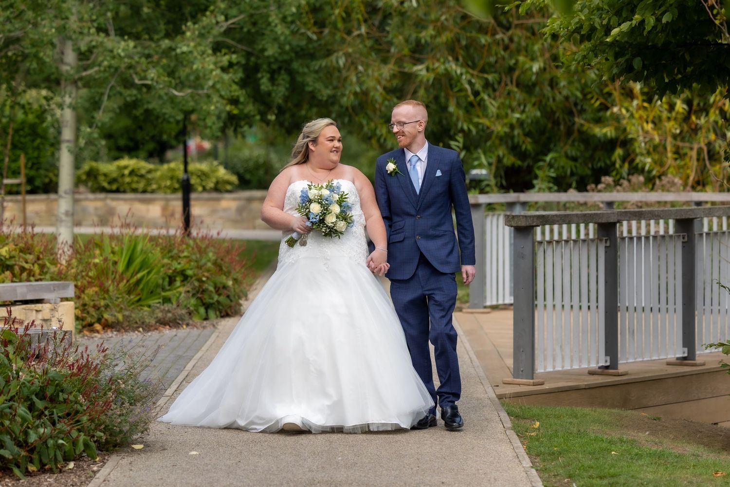 Bride and groom walking together on a garden pathway surrounded by lush greenery and autumn foliage.