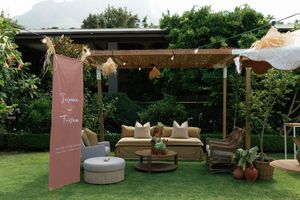 Rustic outdoor lounge area with wooden pergola, string lights, cozy seating and pink welcome sign in garden setting.