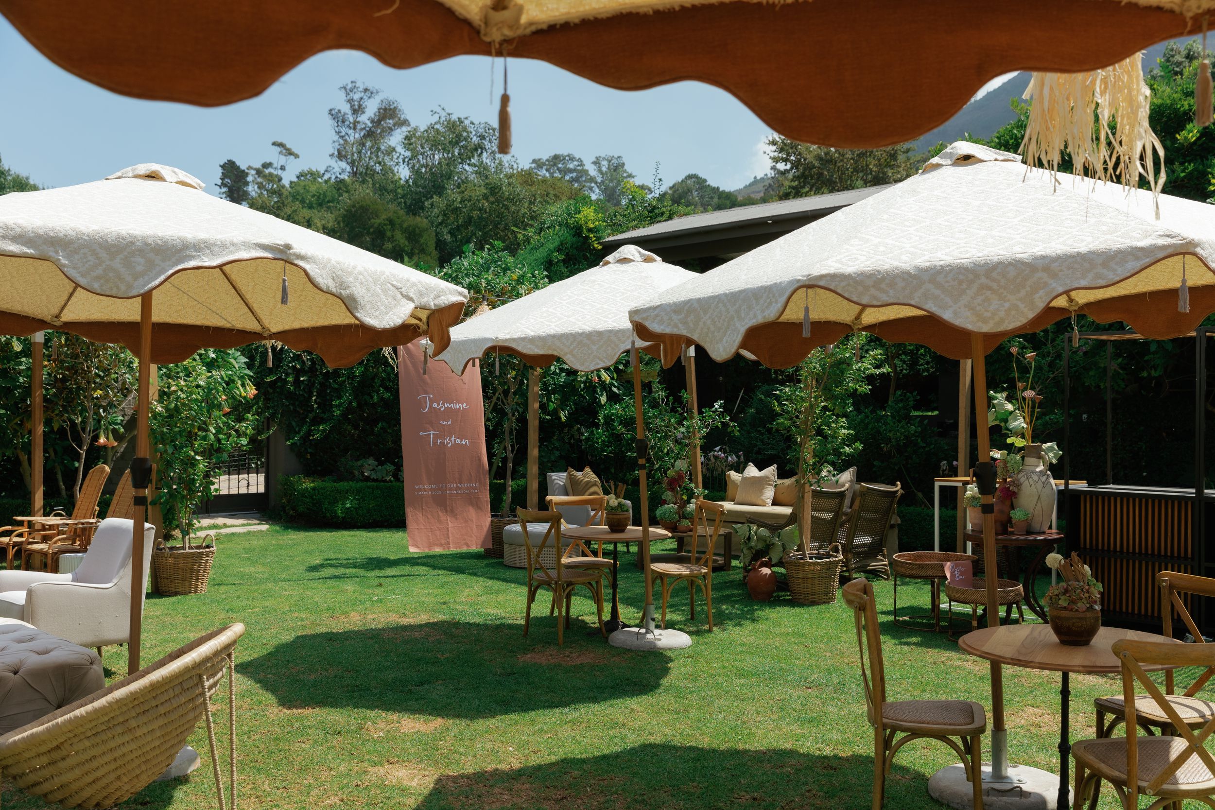Outdoor dining area with white umbrellas and wooden furniture arranged on a grassy lawn near trees.