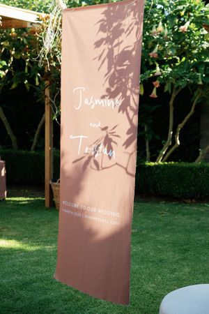 Wedding signage banner with leaf shadows cast on pink background displayed on green lawn.