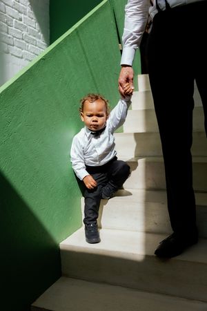 A toddler plays in a sunlit hallway with green walls while an adult figure stands nearby in shadow.