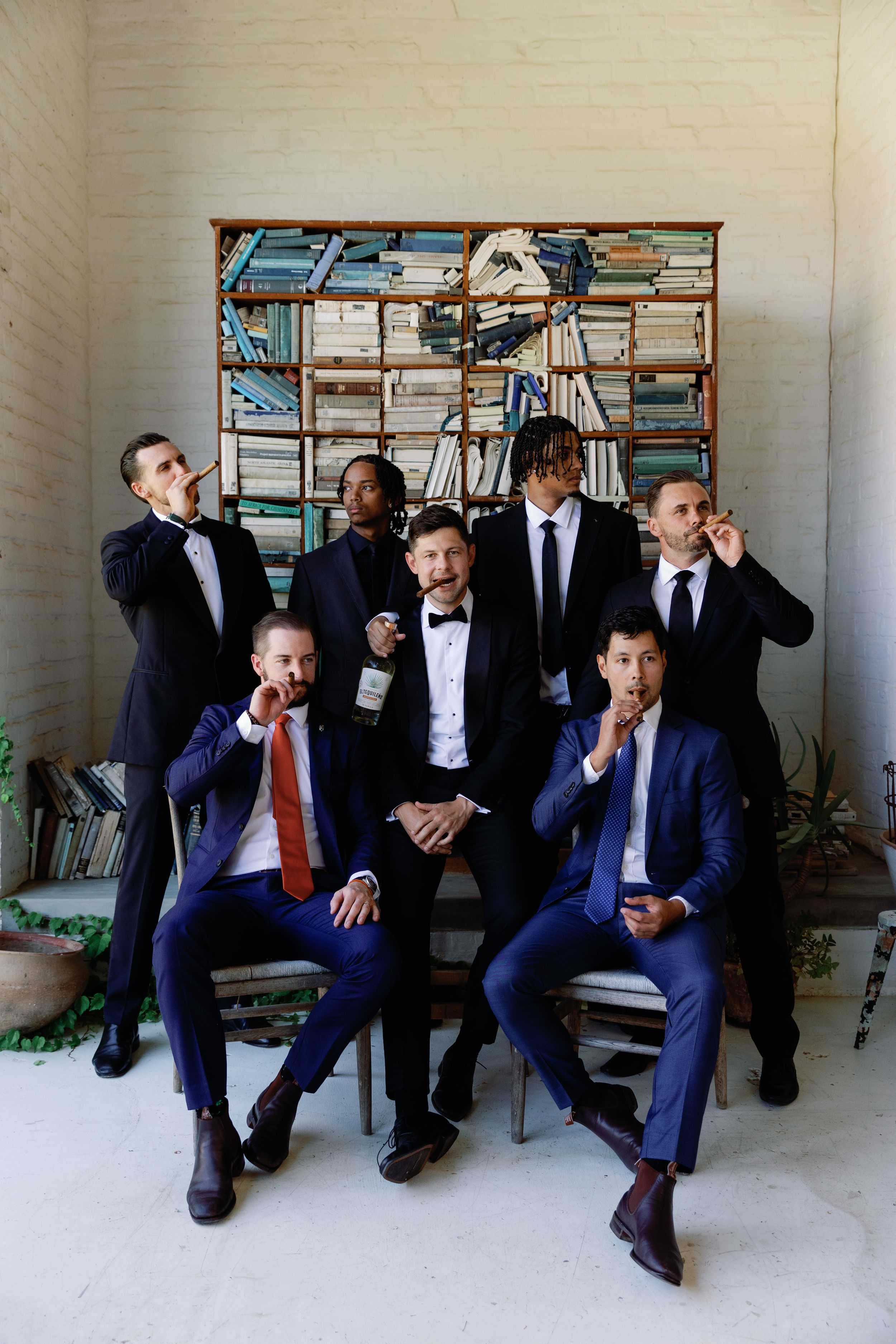A group in formal suits strike playful poses against a bookshelf backdrop during a wedding celebration.