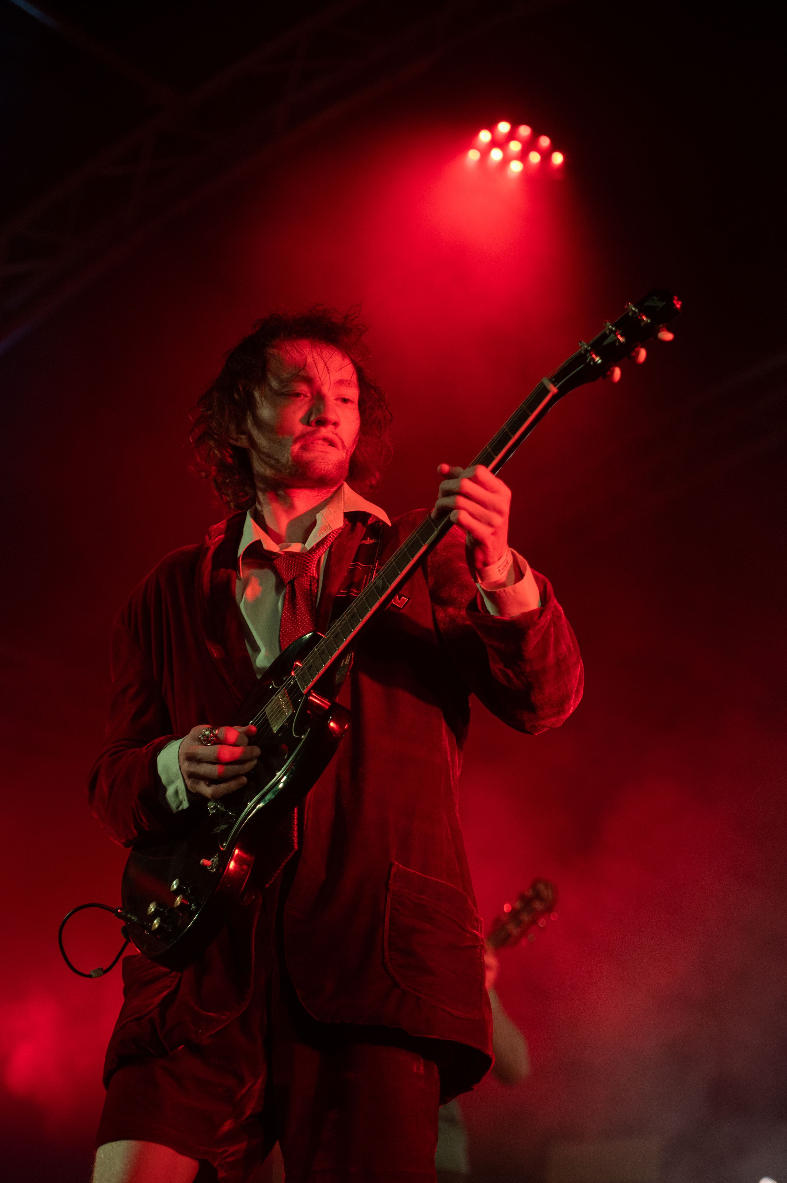 Musician performs on stage bathed in dramatic red lighting while playing guitar during an intense concert performance.