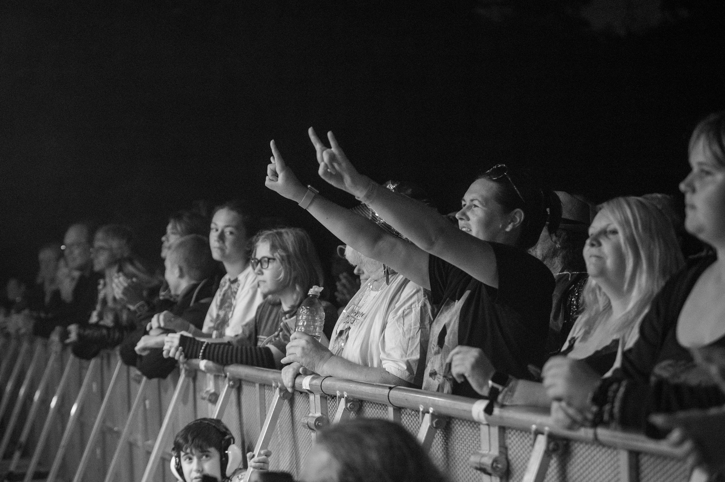 Black and white photo of concert crowd with raised hands at barrier watching performance from front row.