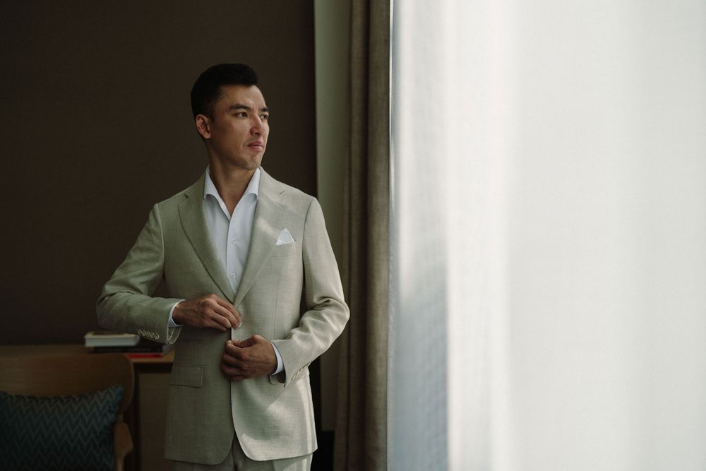 A person in a white blazer sits by a window in a contemplative pose during a photoshoot.