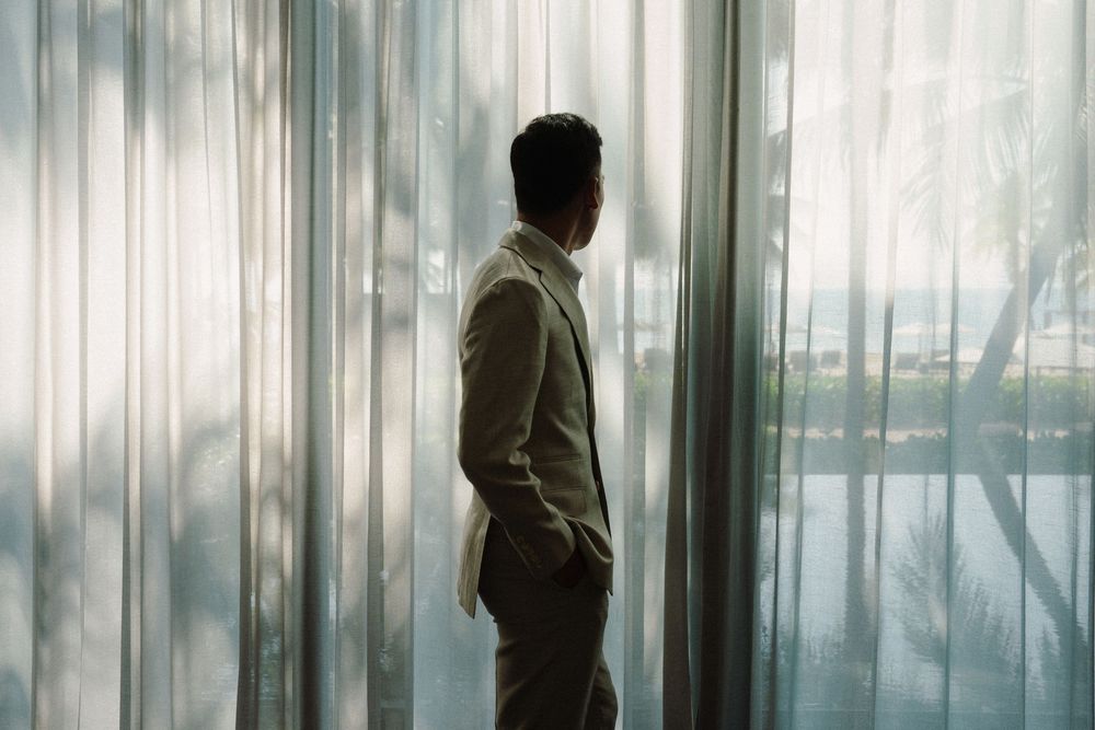 A silhouetted figure stands contemplatively by large curtained windows in a bright, minimalist interior space.