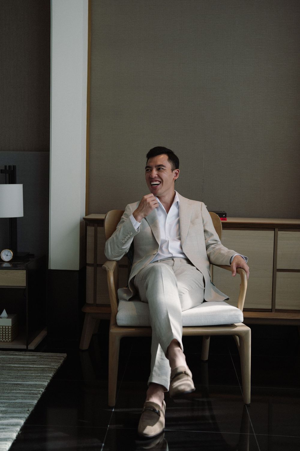 A person in a white suit reclines in a wooden chair while laughing and relaxing in a dimly lit room.