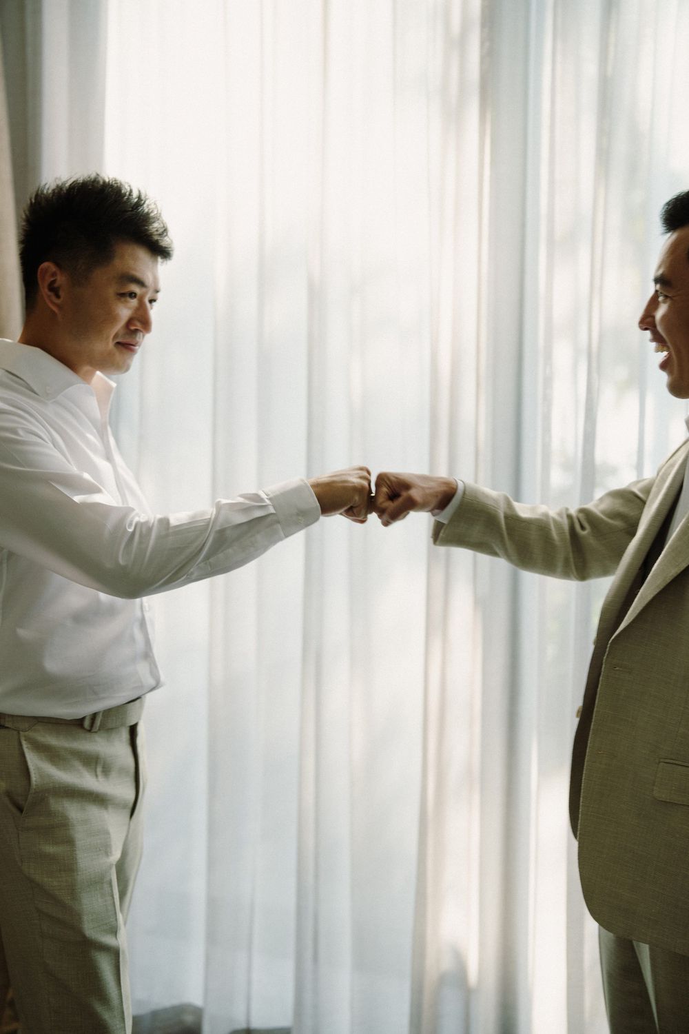 Two people in formal attire fist bump in front of a white curtained window.