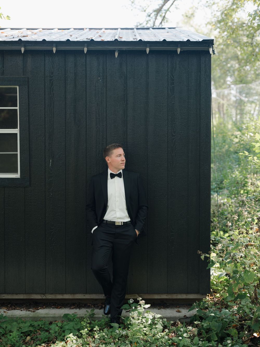 Artistic wedding photo series showing a groom in a black tuxedo against a dark wooden wall in a garden setting.