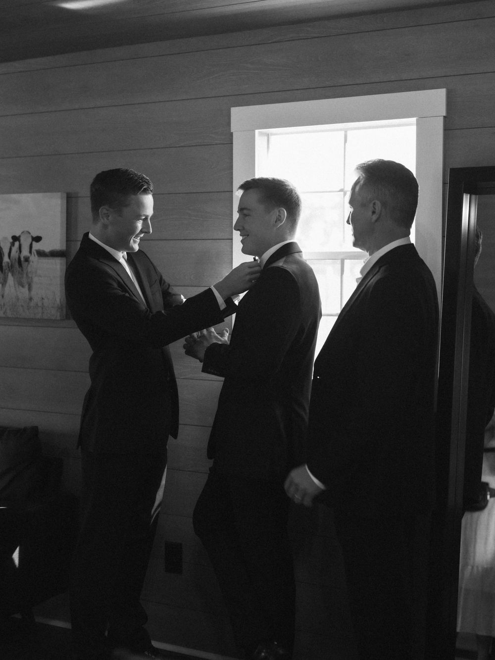 Three men in dark suits stand in a room, with one adjusting another's tie or collar in this black and white photograph.