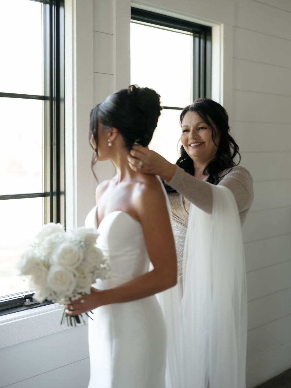 A tender moment between a bride and groom sharing embraces near a bright window with white bouquet.