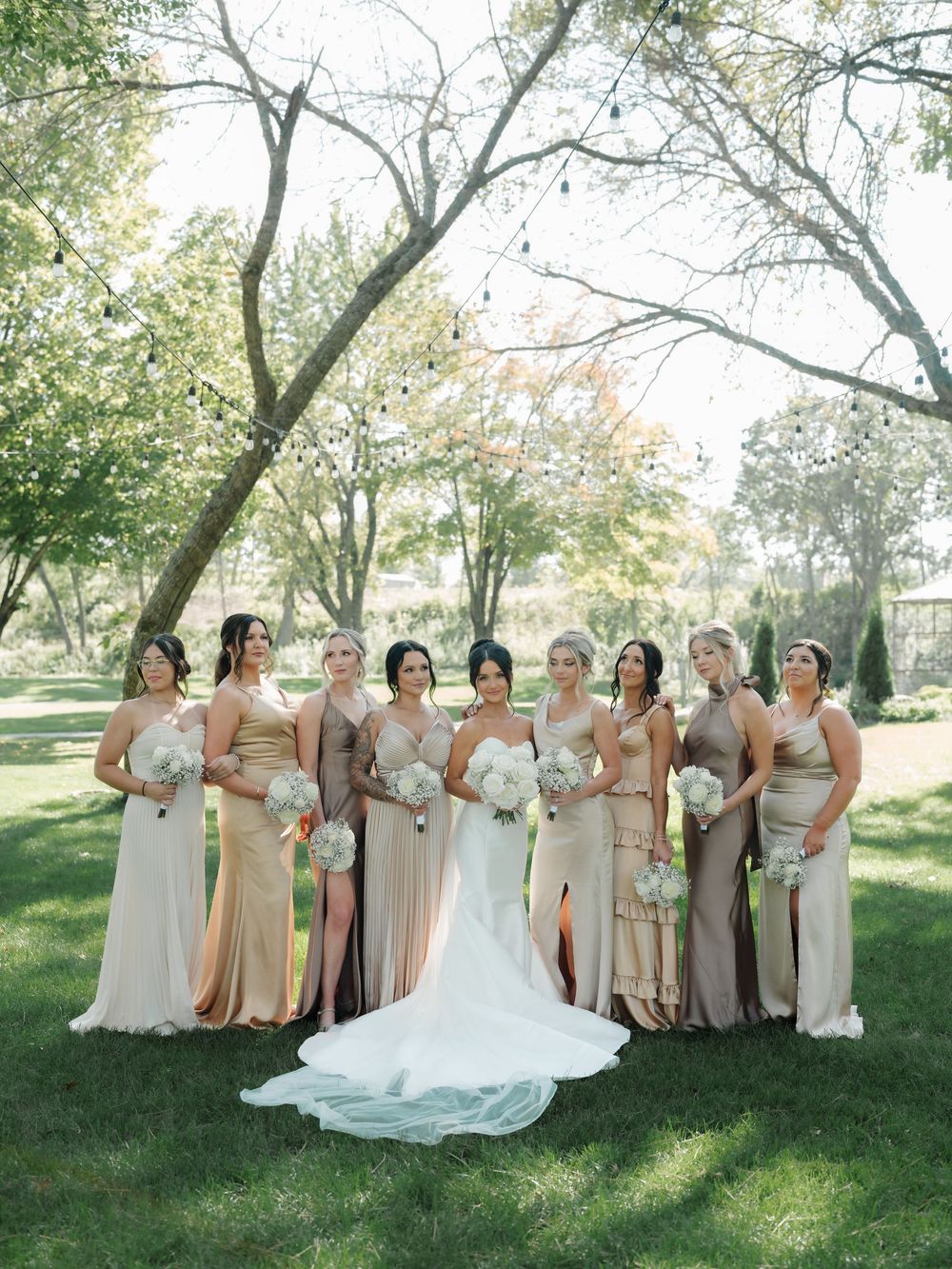 Wedding party in neutral-colored dresses poses together on a grassy lawn under trees.
