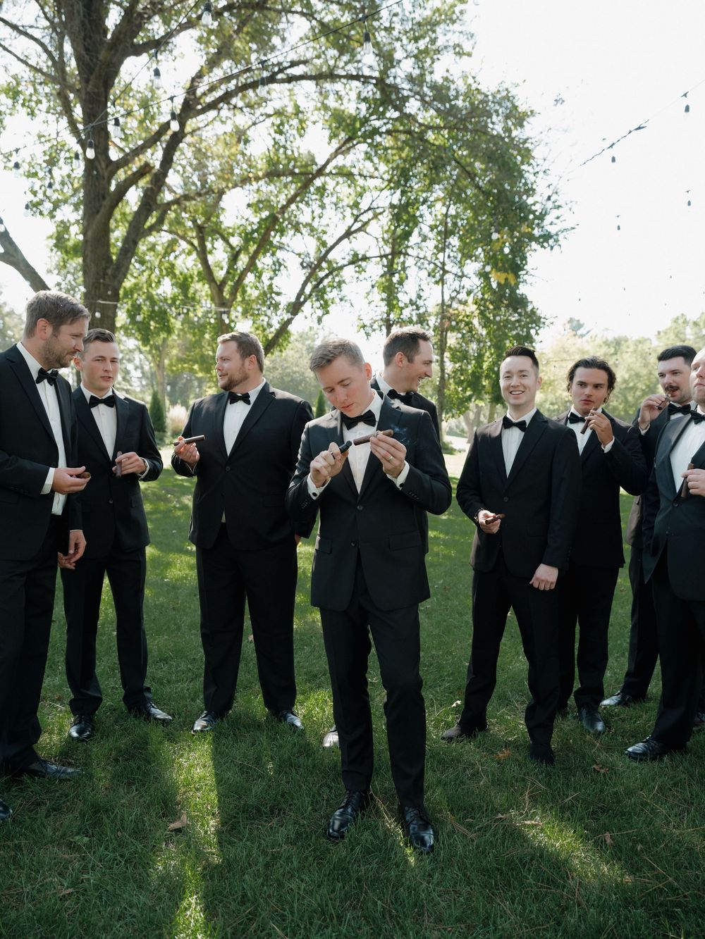 Groomsmen in black tuxedos standing in a line outdoors sharing drinks and laughing together at a wedding celebration.