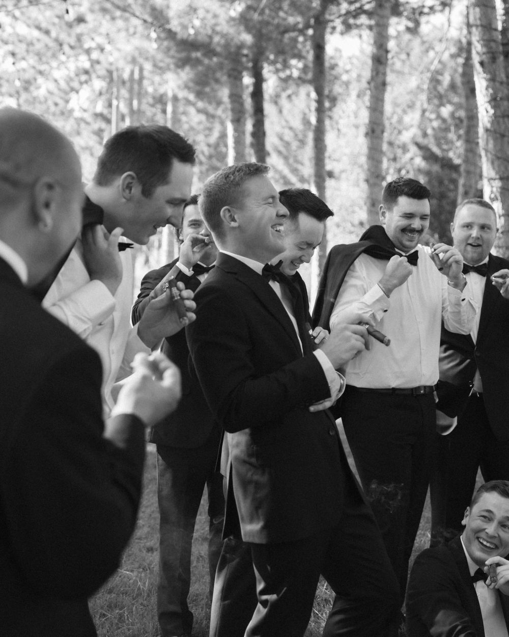 Groomsmen in black suits share a candid moment of laughter in a wooded outdoor setting.