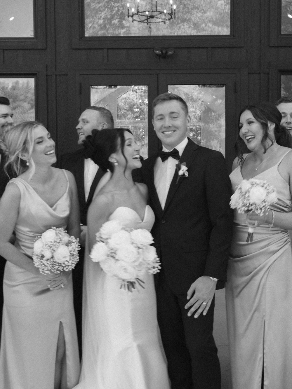 Wedding party poses together against dark wall with bridesmaids in light dresses holding bouquets.