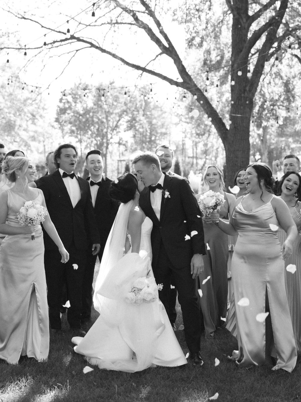 Wedding party dressed in formal attire jumps in unison for a playful outdoor group photo under trees.