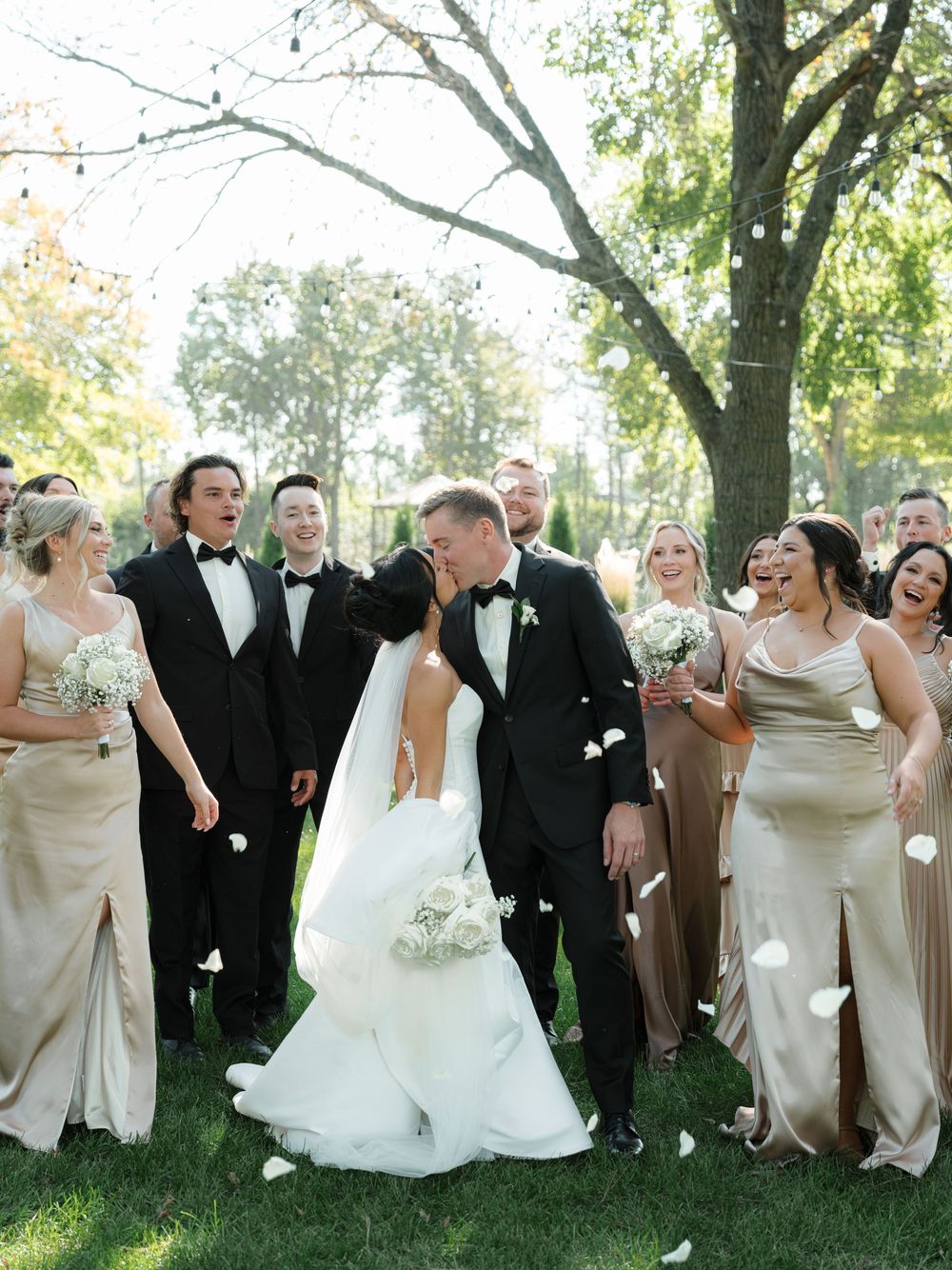 A wedding party laughs together while posing outdoors under trees in formal attire.