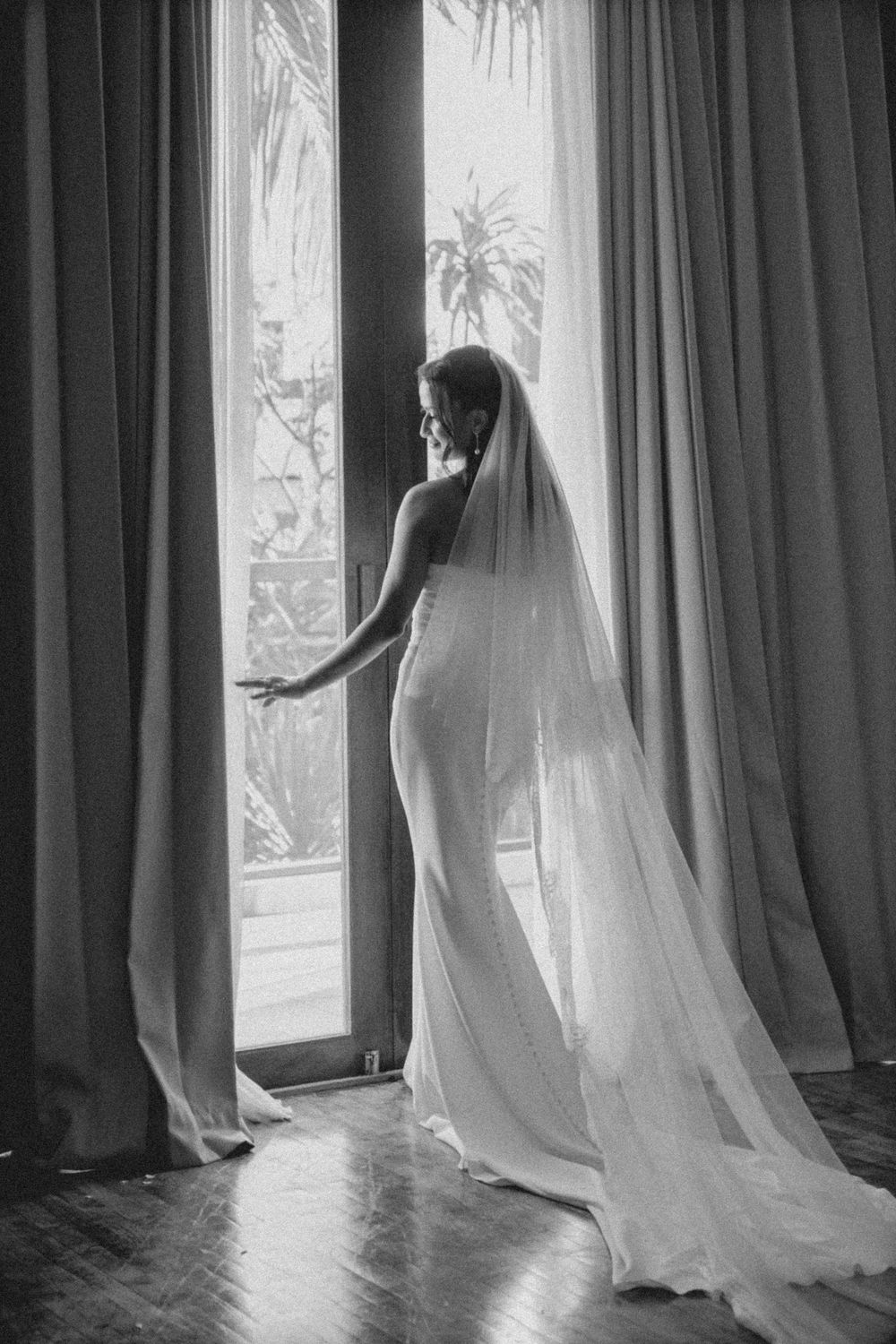 A silhouetted figure in a wedding gown and flowing veil stands elegantly by a sunlit window with palm trees outside.