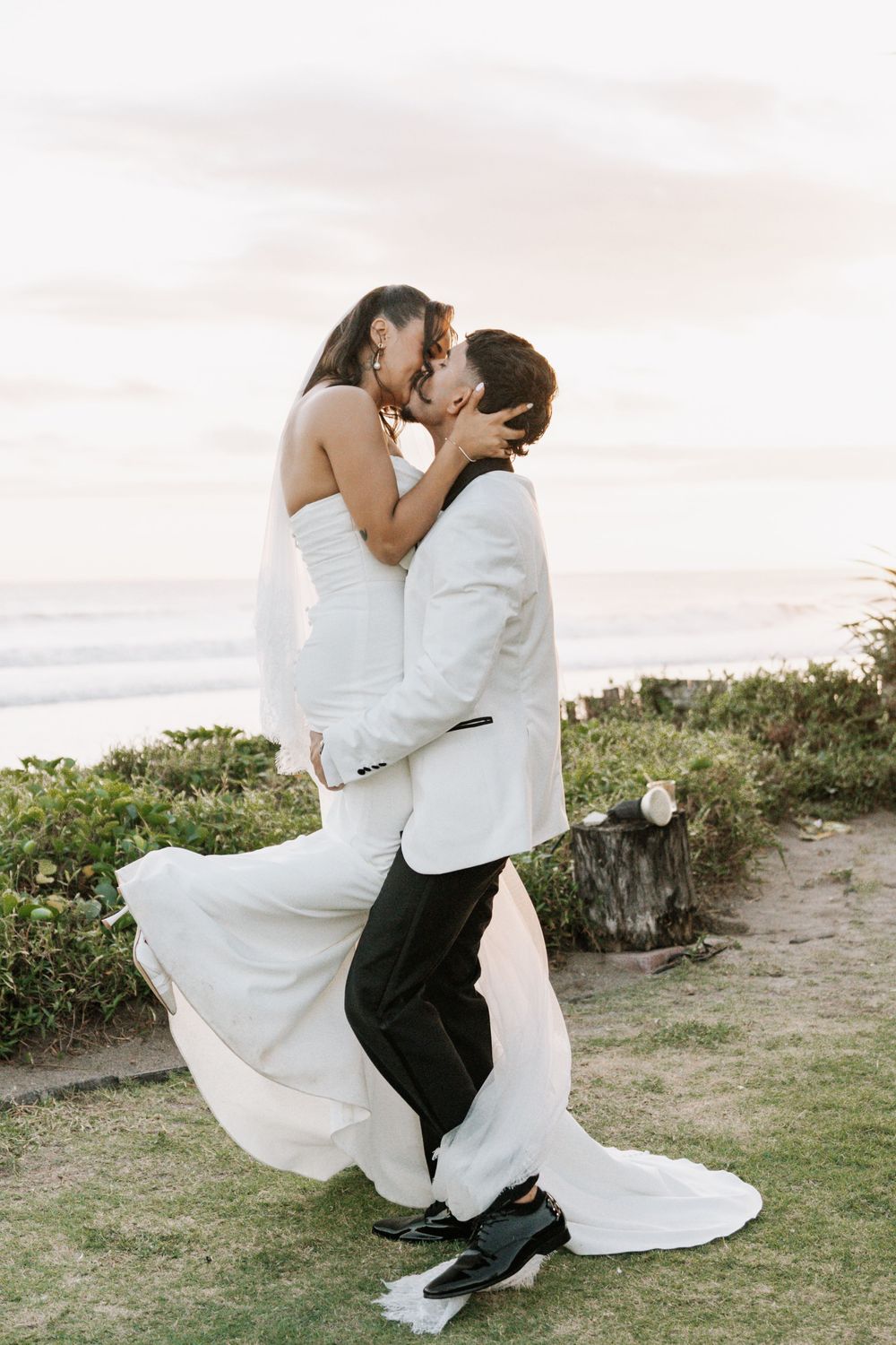 Romantic wedding couple embraces and kisses on grassy coastal cliff during sunset in matching white attire.