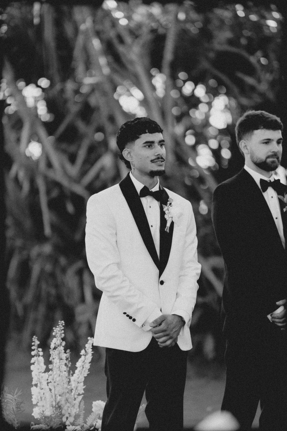 Groomsmen in black and white formal attire stand together during an outdoor evening wedding ceremony.