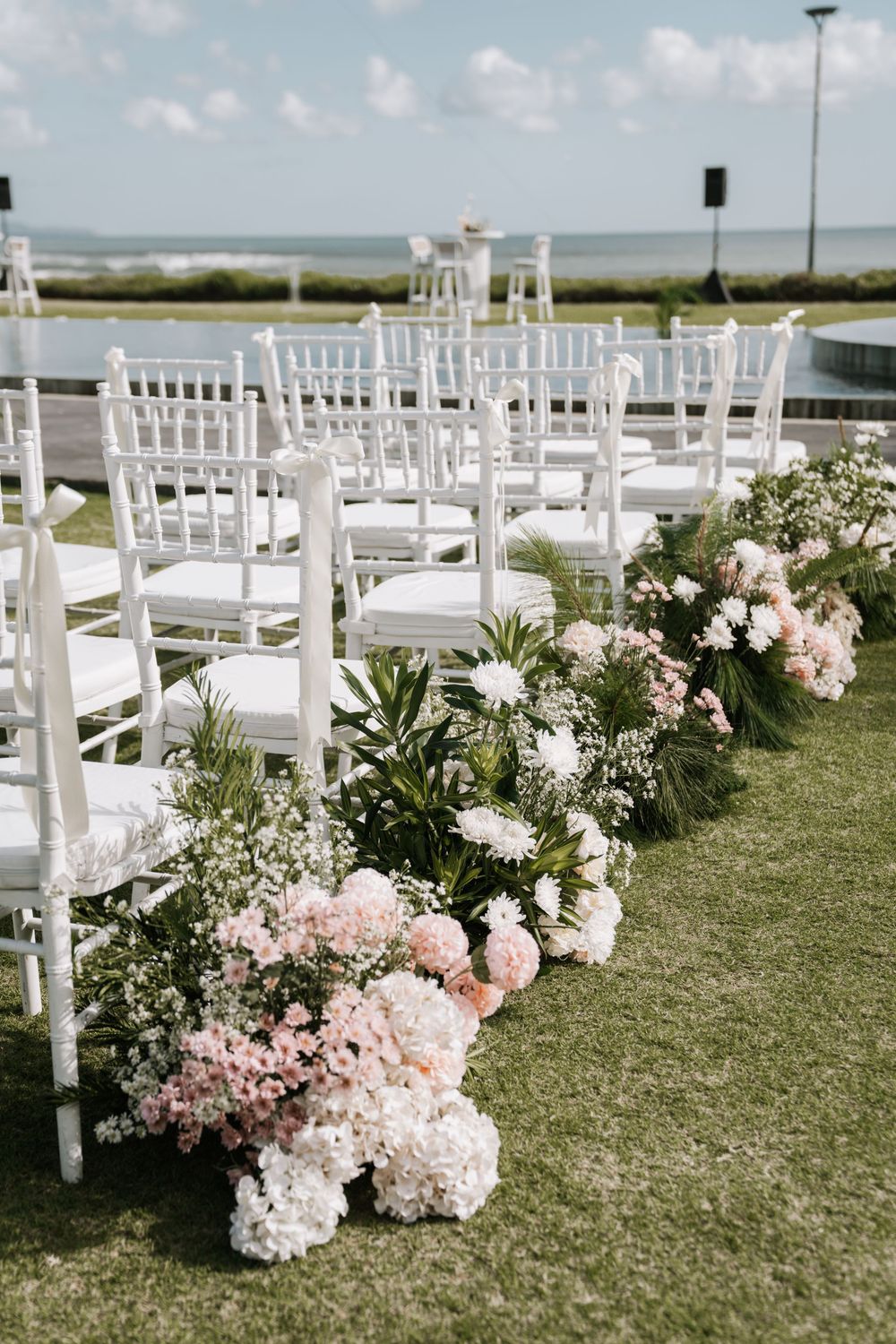 White garden chairs and delicate pink floral arrangements line an outdoor wedding ceremony aisle by the ocean.