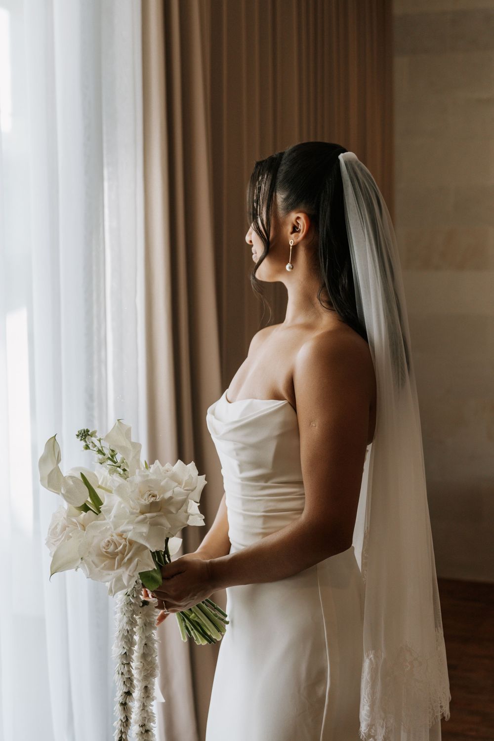 Bride in elegant white strapless gown holds white floral bouquet while gazing out window in soft natural light.