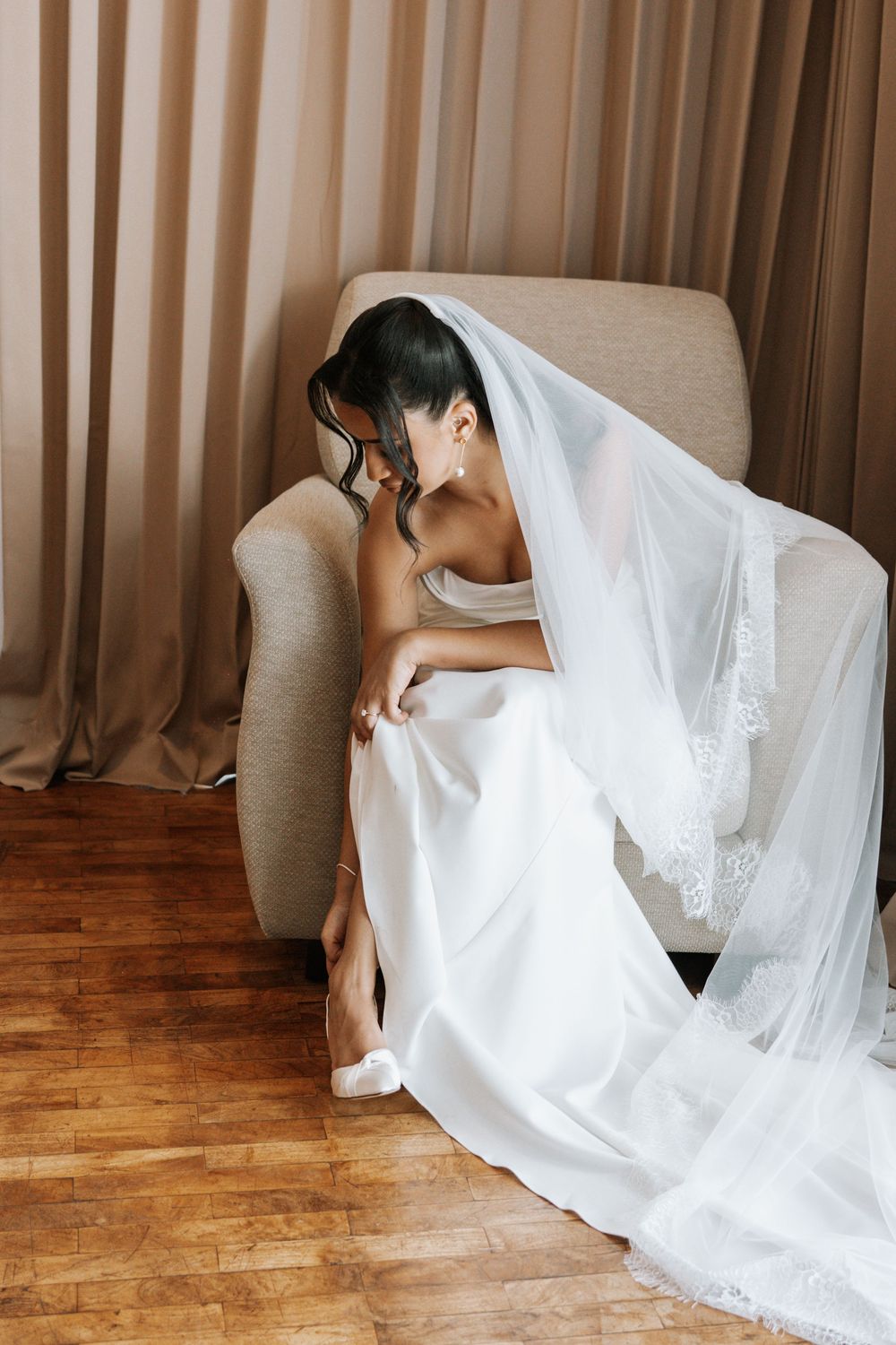 A bride in a flowing white wedding dress sits gracefully on a hardwood floor during an indoor photo session.