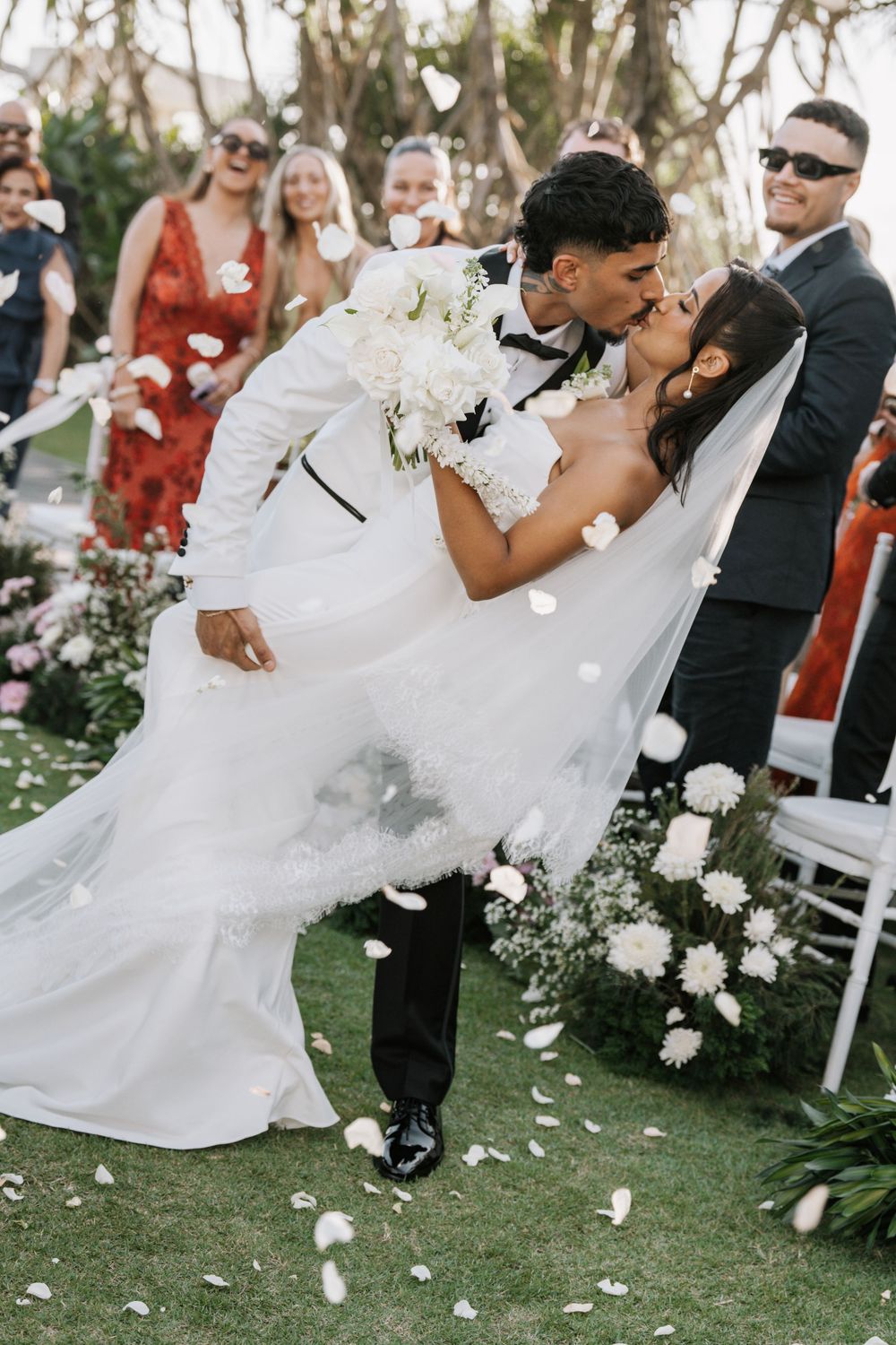 Wedding couple performs a romantic dip pose on grass surrounded by flower petals and guests.