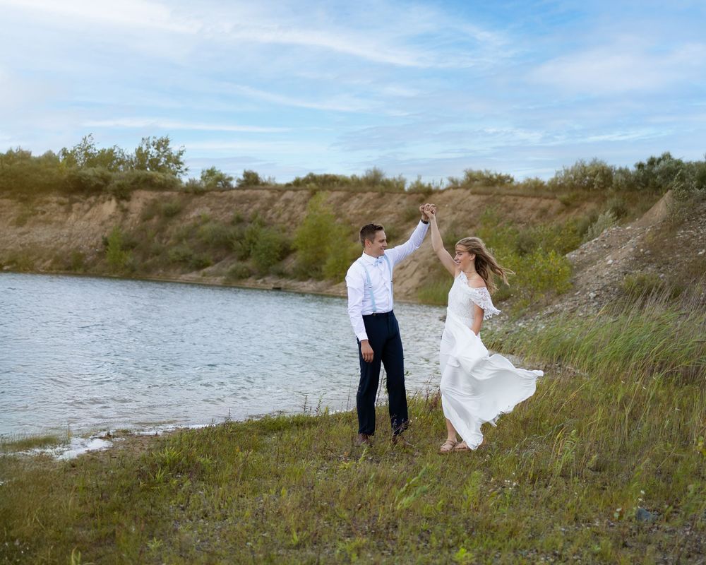 A couple in wedding attire dance joyfully together on a grassy hillside near a serene lake on their special day.