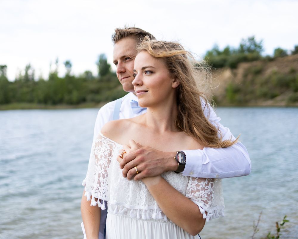 A couple in white attire dances together by a serene lakeside on a sunny afternoon.