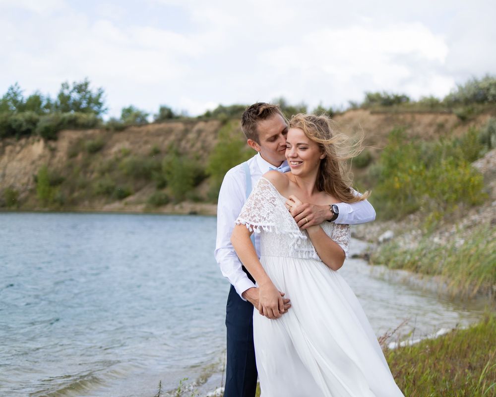 Two people share a romantic embrace in a white wedding dress and navy suit along a lakeside shore with gentle waves.