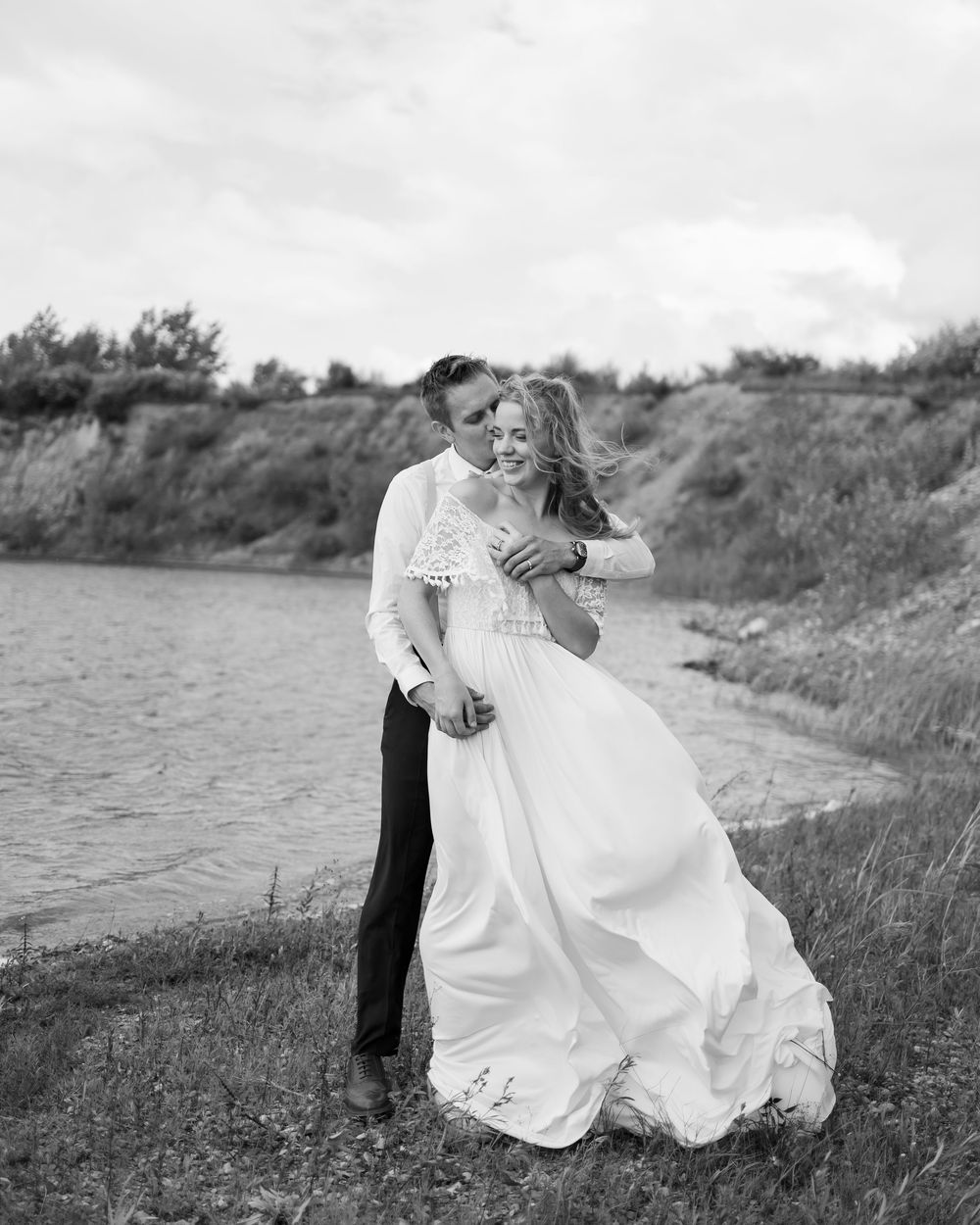 A romantic black and white portrait of a couple embracing by a riverbank, with the bride's white dress flowing in the wind.