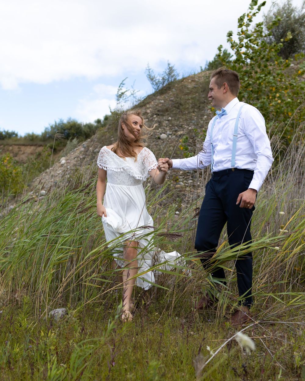 A couple in summer attire stands among tall grass on a hillside during a sunny afternoon.