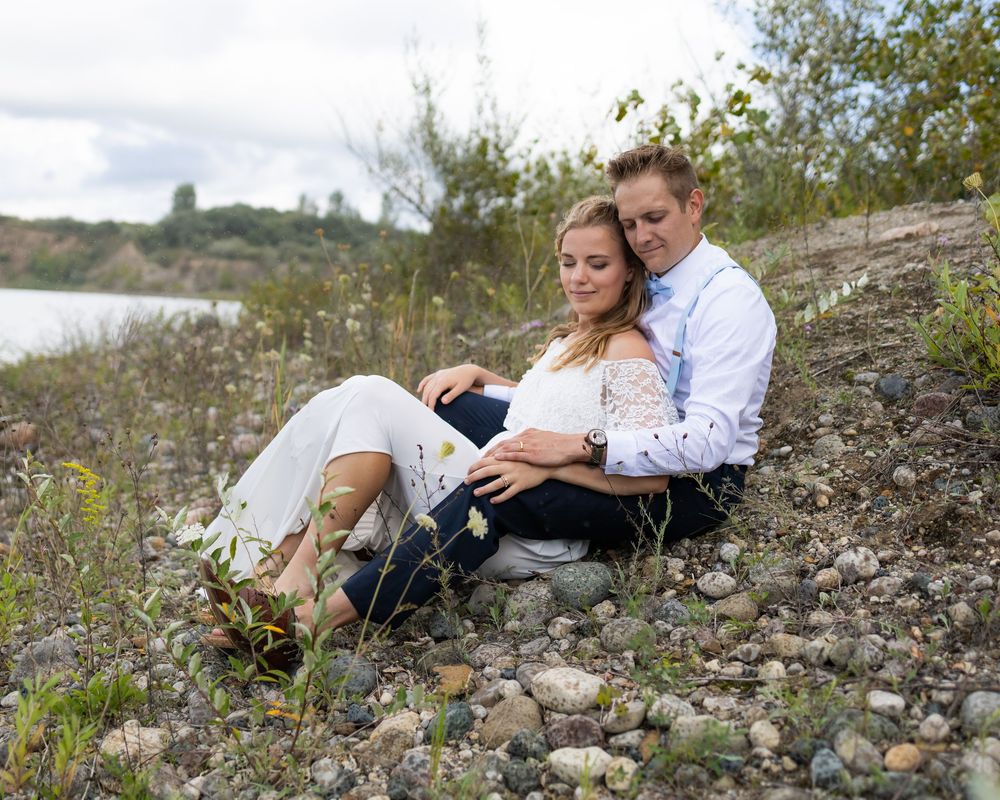 A couple sits together on a rocky riverbank surrounded by natural vegetation on a sunny day.