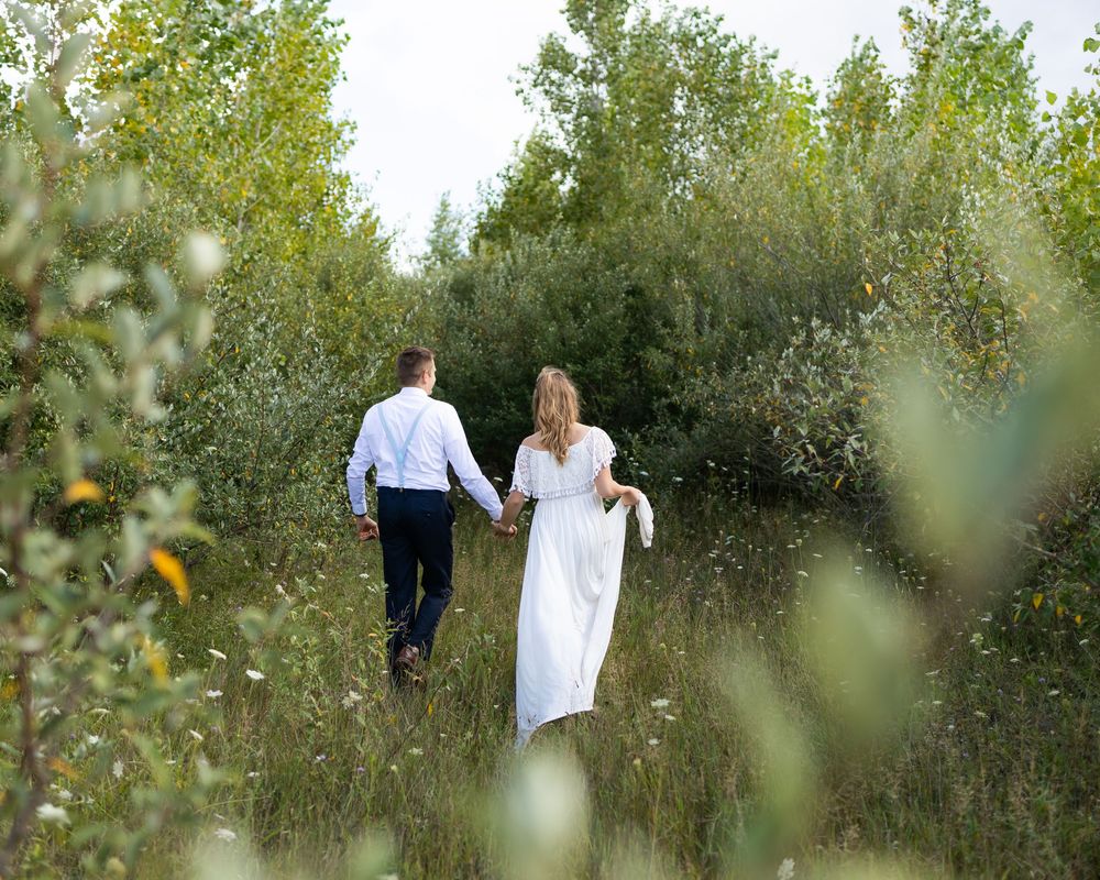 A couple walks through a lush green pathway surrounded by tall trees and wild grass on a sunny afternoon.