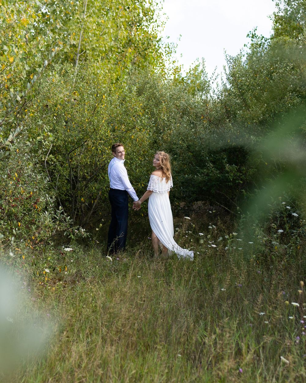 A couple in wedding attire stands together in a natural setting with tall grass and yellow-leaved trees.