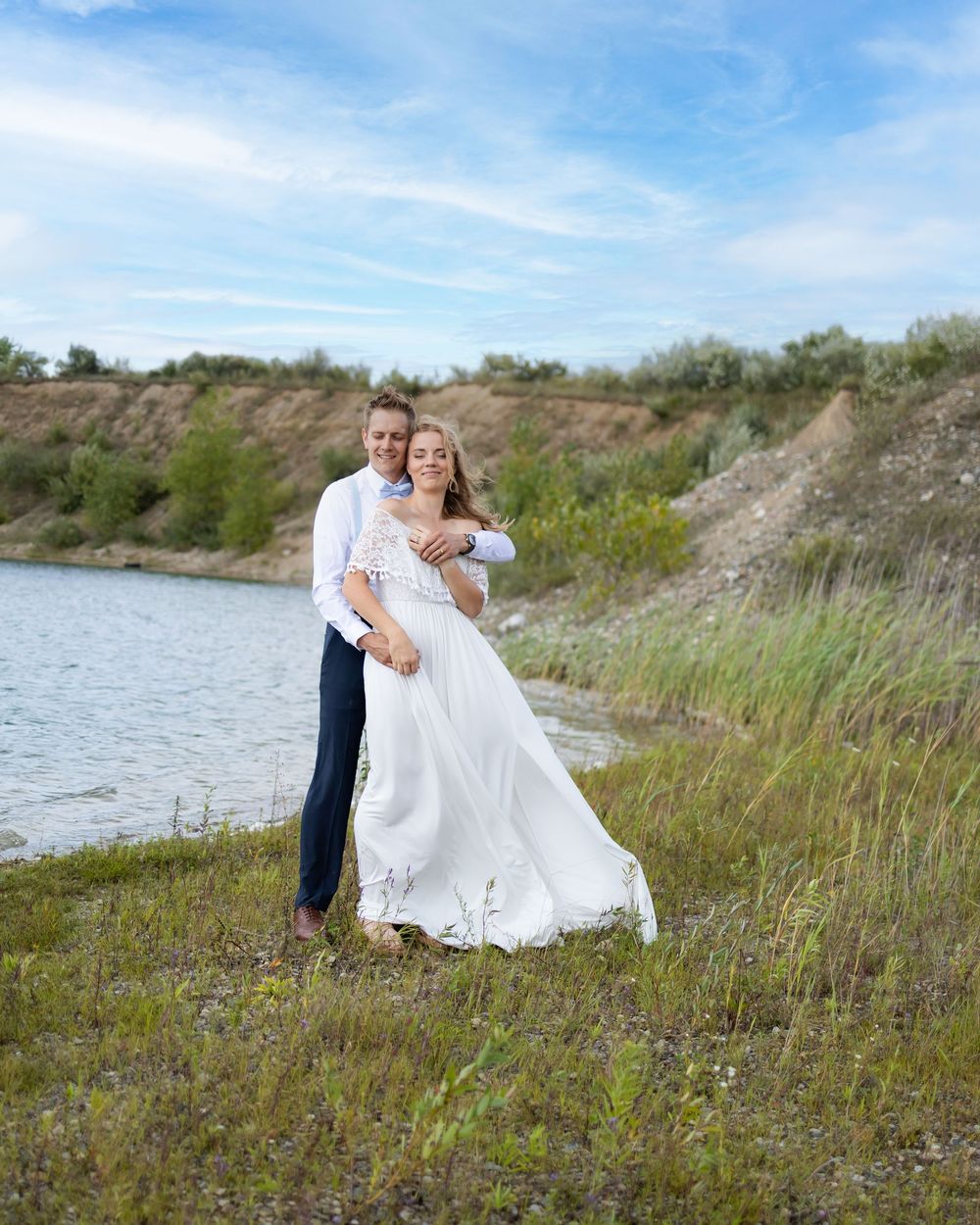 A couple in wedding attire embrace on a grassy hillside near a lake on a beautiful sunny day.