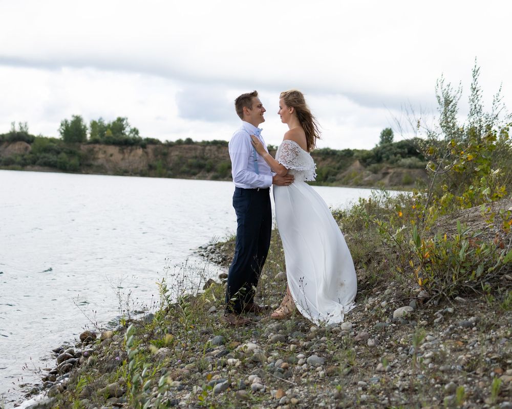 A couple in formal wedding attire embraces on a lakeside slope with cloudy skies above.