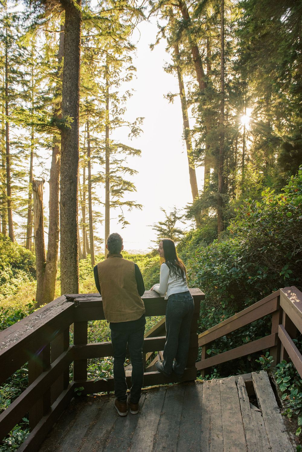 Two people stand on a wooden deck viewing platform in a sunlit forest with tall evergreen trees at sunset.