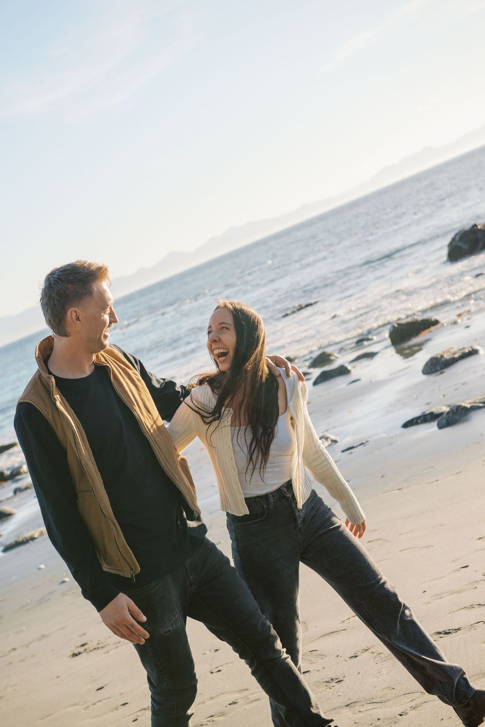 A couple shares joyful laughter while walking together on a rocky beach at sunset.