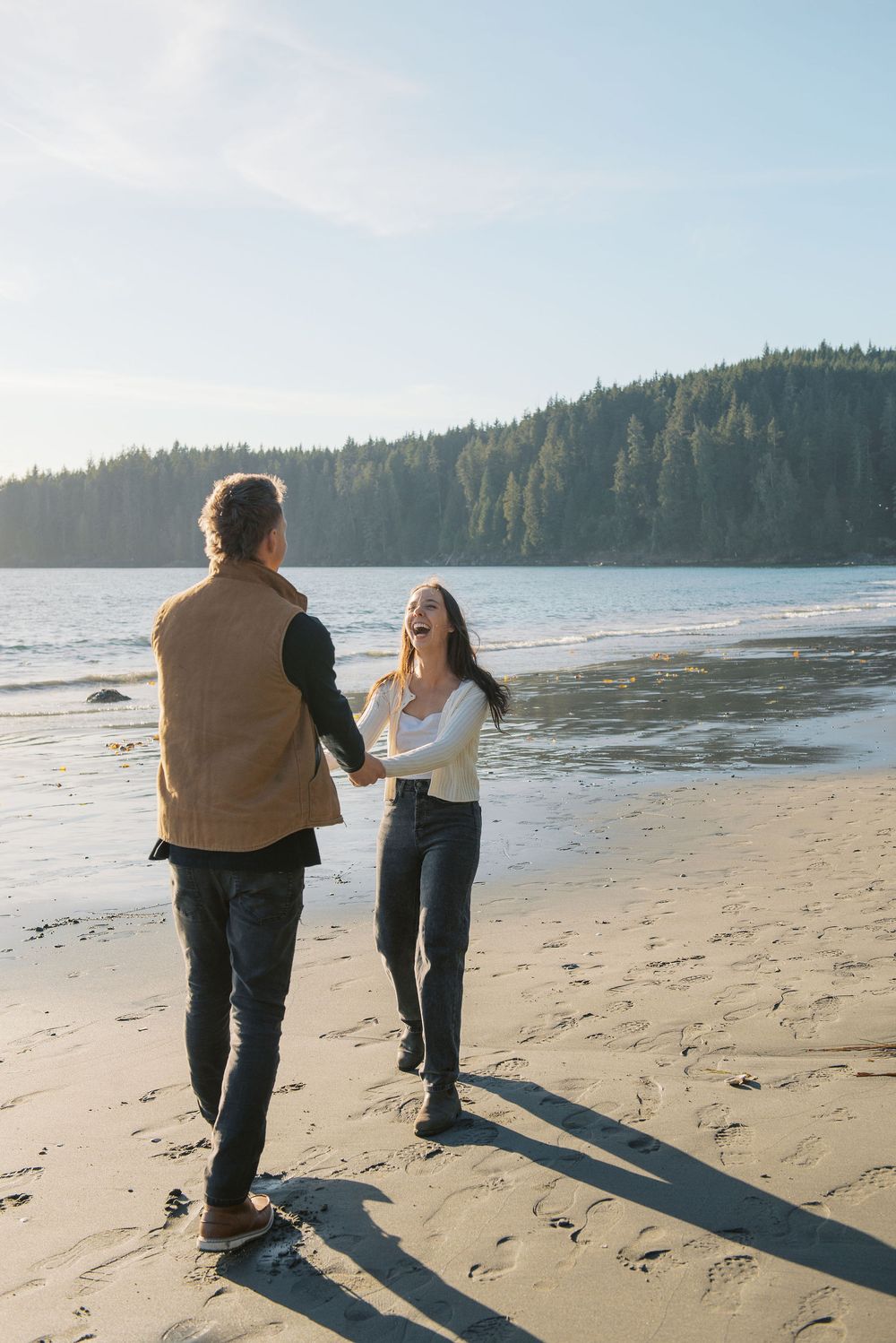 Two people walk together on a scenic beach with evergreen trees and ocean waves in the background.