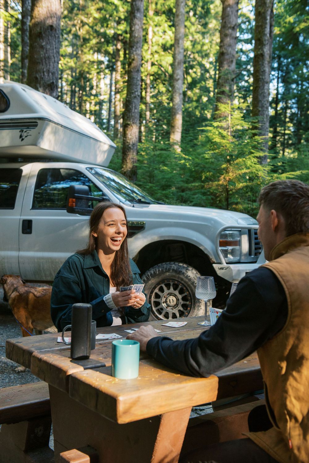Two people enjoying coffee at a wooden picnic table next to their camper van in a forest campsite.