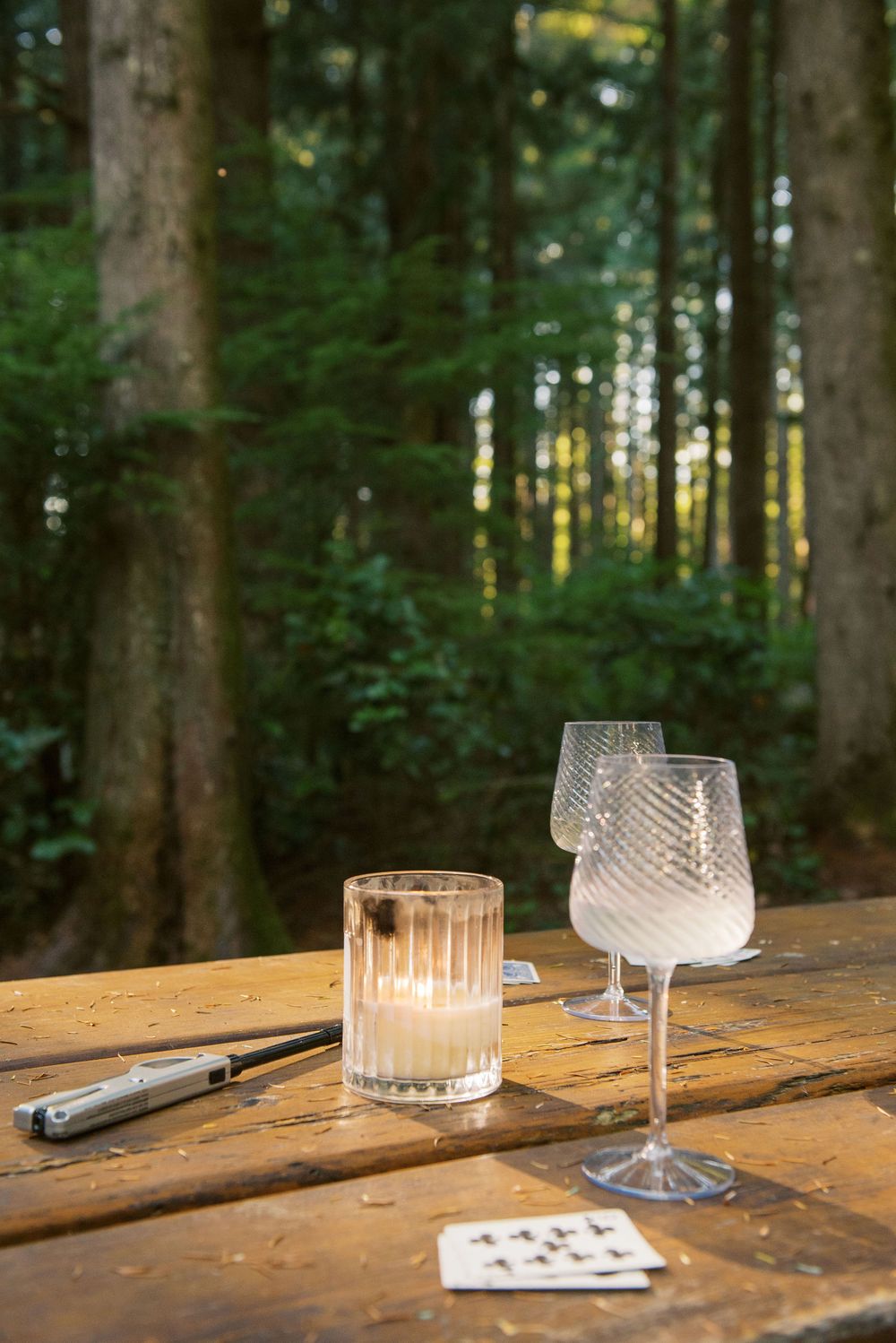 Playing cards and wine glasses sit on a rustic wooden table in an outdoor forest setting at dusk.