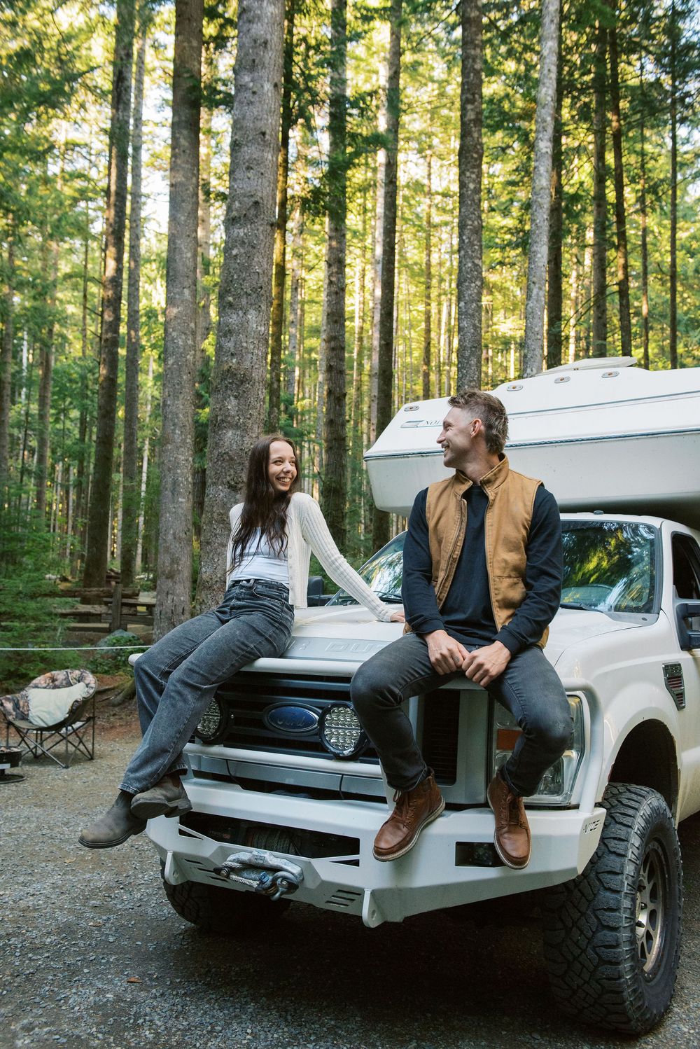 Two people sitting in the back of a white pickup truck at a forest campsite during golden hour.