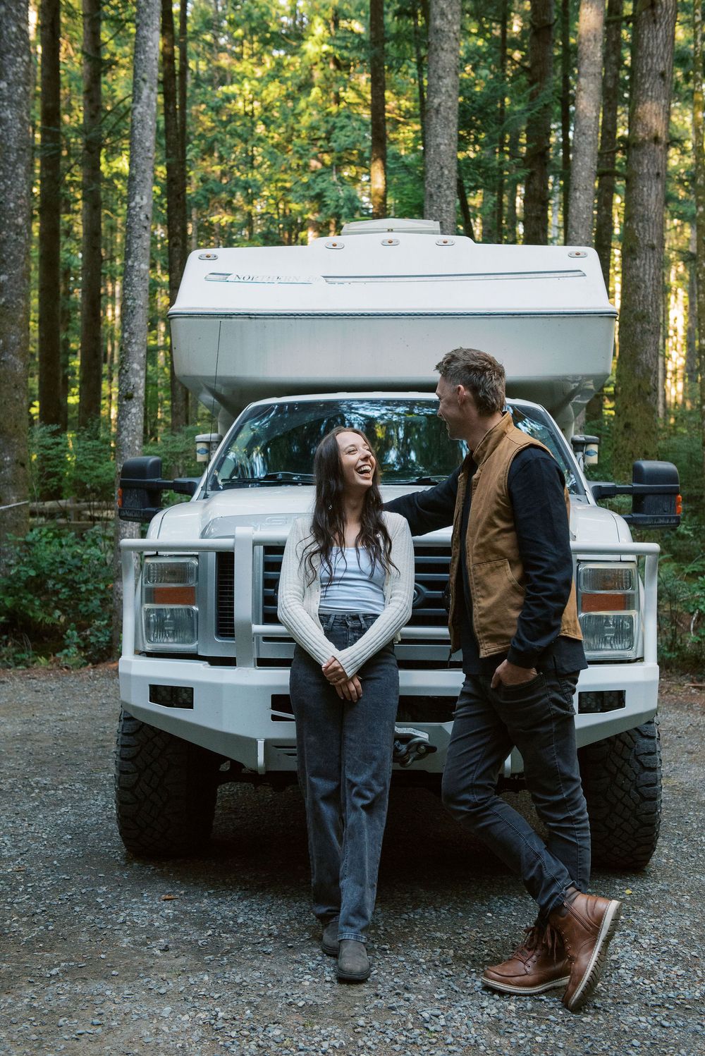 A truck with a camper shell parked in a wooded forest area on a dirt path.