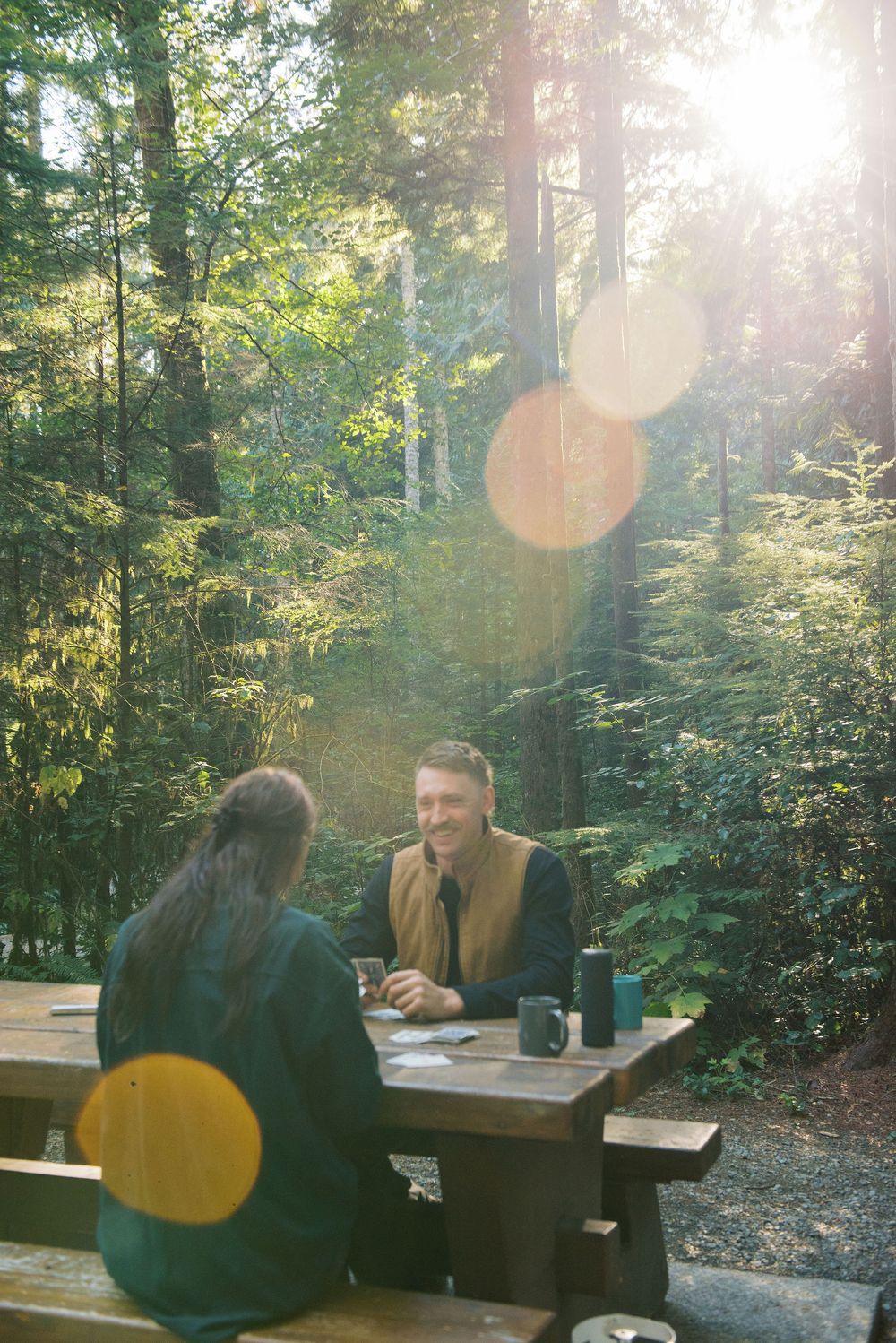 Two people having a conversation at a wooden picnic table in a sunlit forest setting with lens flare effects.