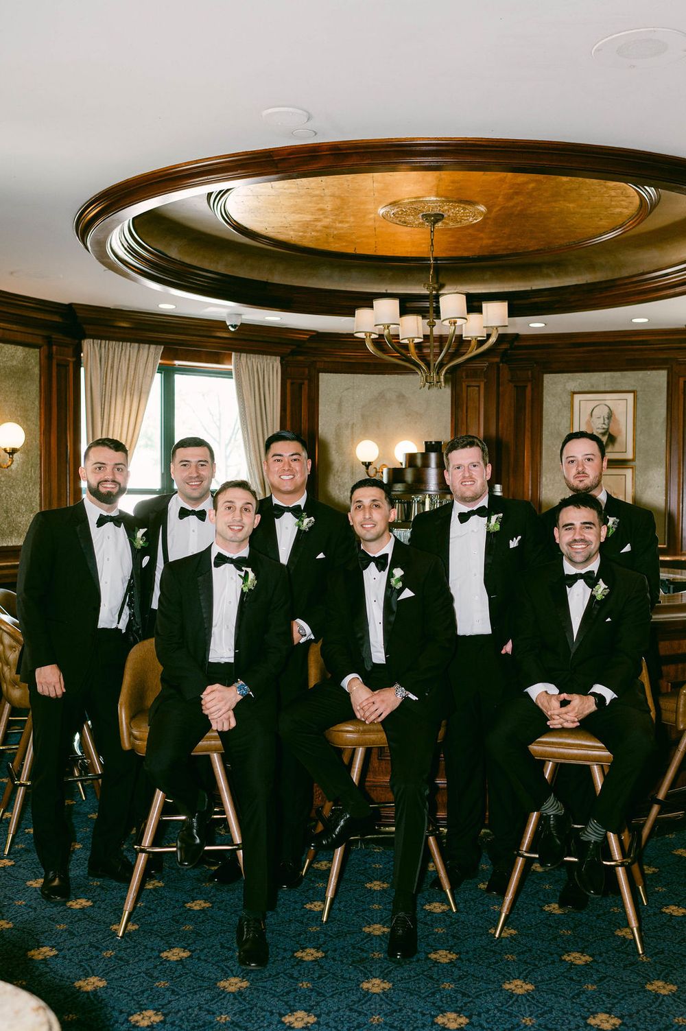 Groomsmen in black tuxedos sit on wooden stools in an elegant room with patterned blue carpet and ornate ceiling at the Willard hotel in DC.