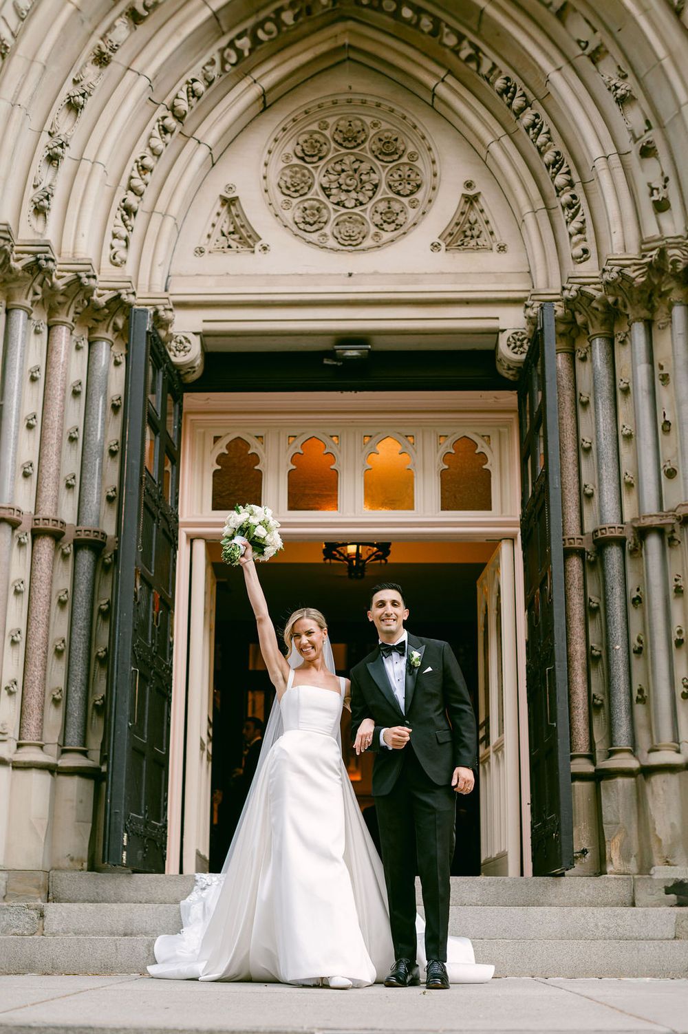 A wedding couple celebrates outside an ornate Gothic church entrance with elegant architectural details.